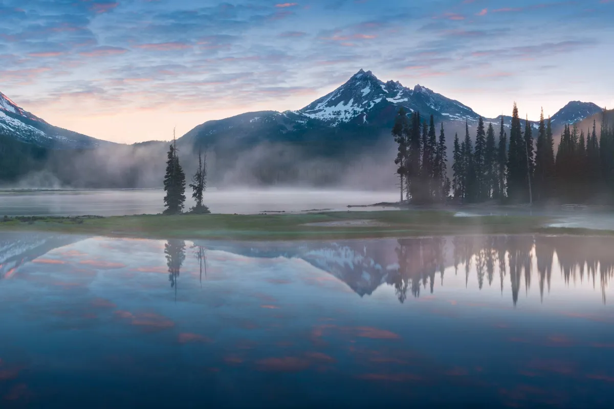A tranquil lake mirrors snow-capped mountains, shrouded in morning mist and framed by tall evergreens, evoking the serenity of the Cascade Lakes and natural beauty near Bend.