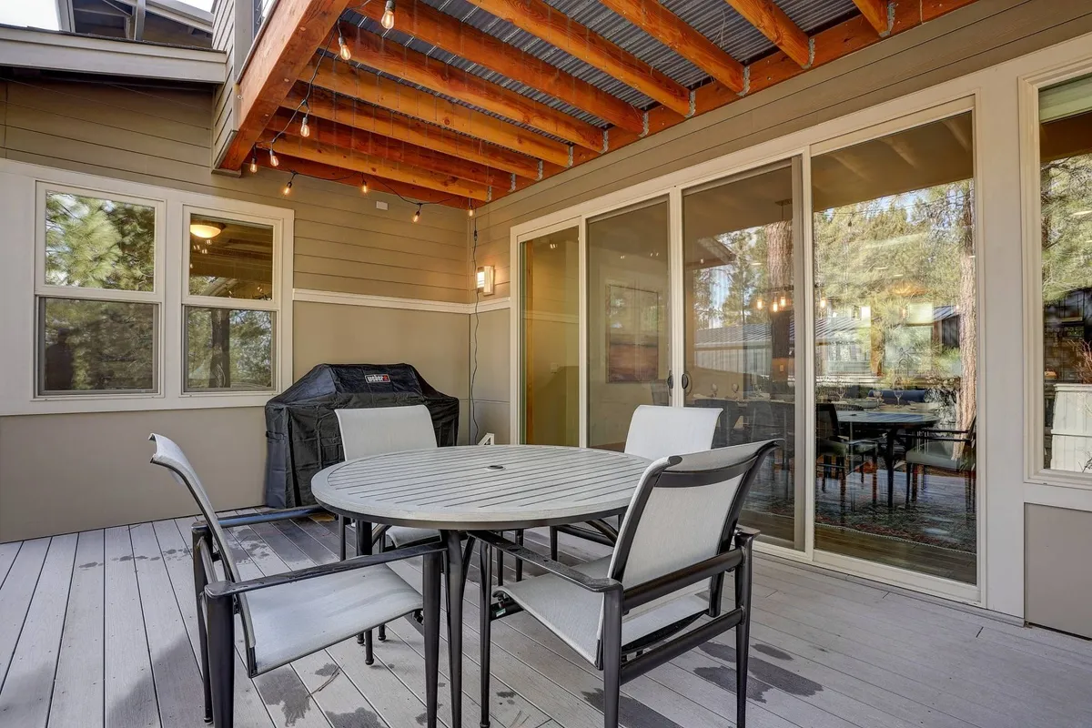 An inviting round dining table sits on a deck with four chairs and a gas grill in the background. Large windows reflect the surrounding trees, emphasizing the strong connection between home and nature. The setup is ideal for barbecues, casual meals, or stargazing under the open sky.
