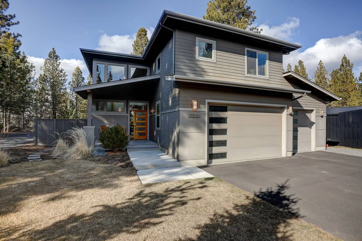 The image showcases the striking exterior of a modern single-family home in Bend, Oregon, featuring clean lines, a warm wooden front door, meticulously landscaped yard, and a spacious driveway. The inviting porch catches the glow of sunset, hinting at evenings spent outdoors and plenty of space for gatherings.