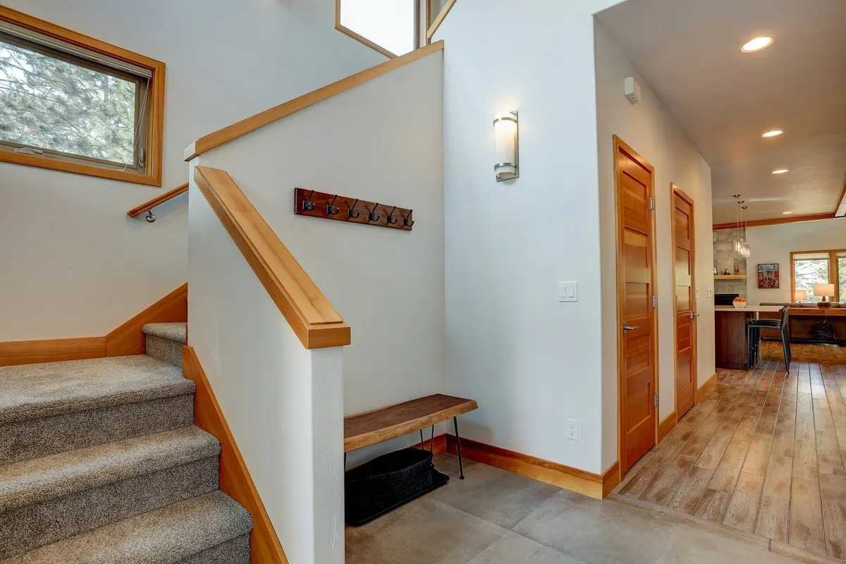 This image captures the home's welcoming entryway, showcasing a staircase, a bench for couples to pause and connect, coat hooks ready for outdoor adventures, and a glimpse of the inviting kitchen beyond. Natural light and functional design highlight the ease of transitions between indoor comfort and exploring Bend.