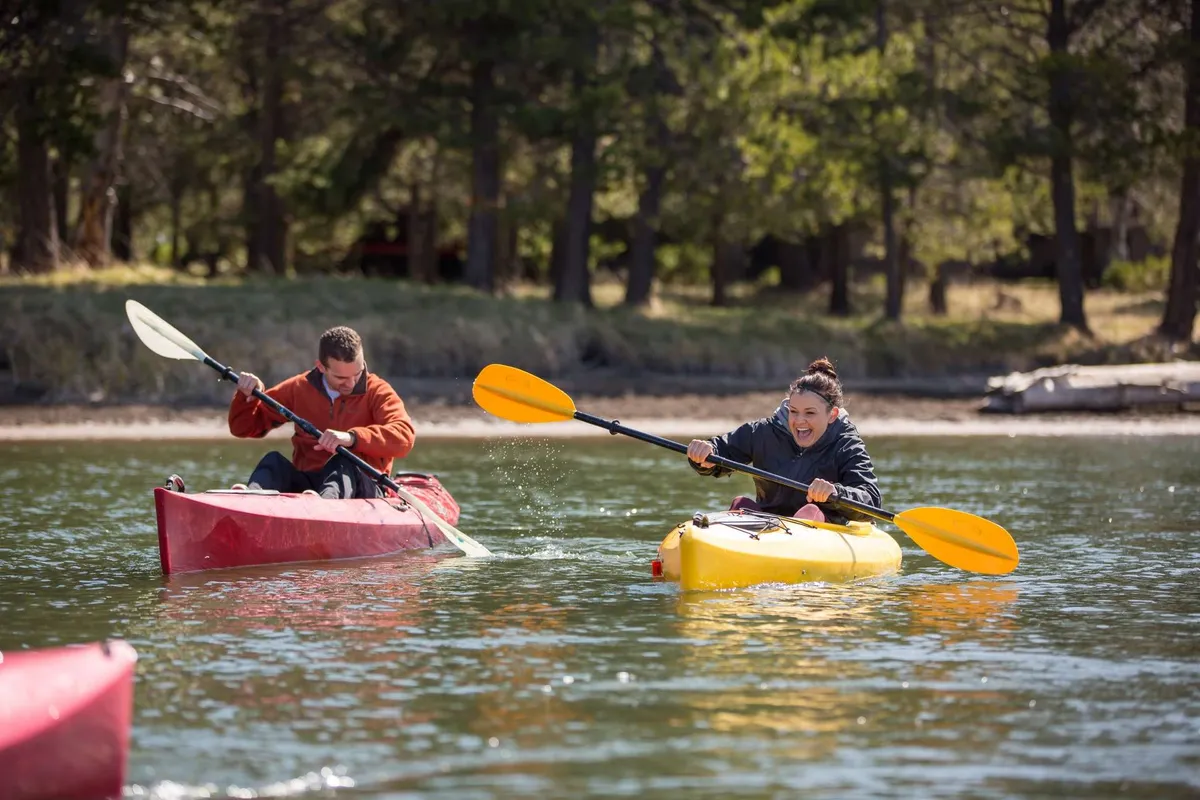 Two kayakers paddle colorful kayaks along a river surrounded by lush greenery. The scene captures active exploration, bright colors, and the natural beauty of the Bend area, suggesting a fun outdoor atmosphere perfect for families or groups.