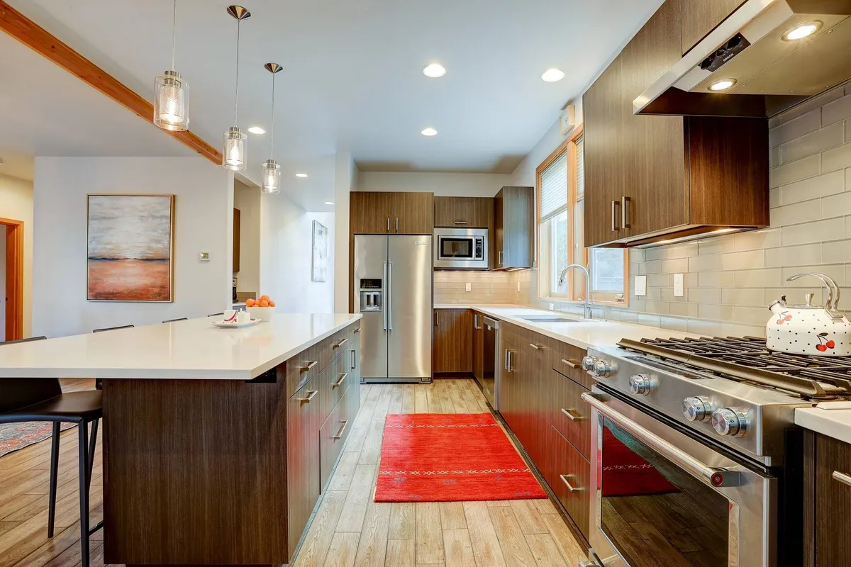 A warm, inviting kitchen featuring an island with seating, stainless steel appliances, a bold red area rug, and modern cabinetry. The layout encourages both cooking and casual interaction among guests or family members.