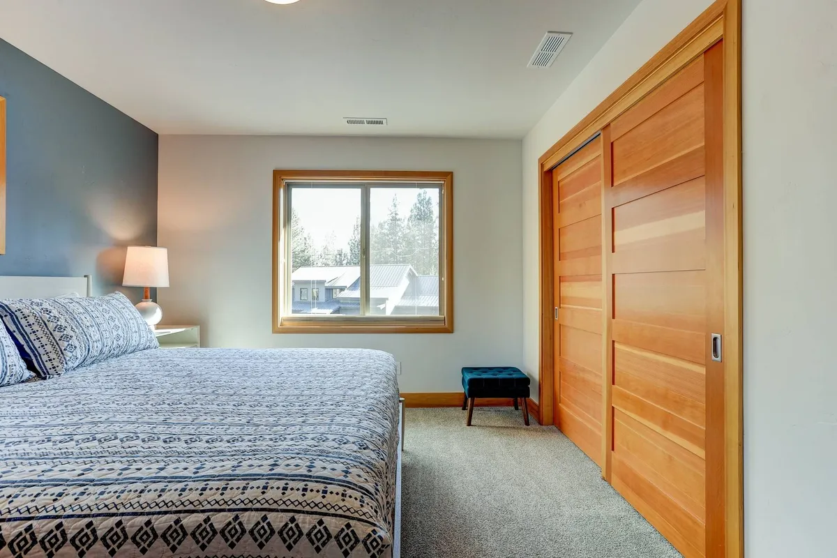 A tranquil and cozy bedroom featuring a bed with patterned bedding, a window with scenic views, a nightstand, lamp, blue accent chair, and sliding closet doors. The space invites relaxation, reading, or quiet mornings.