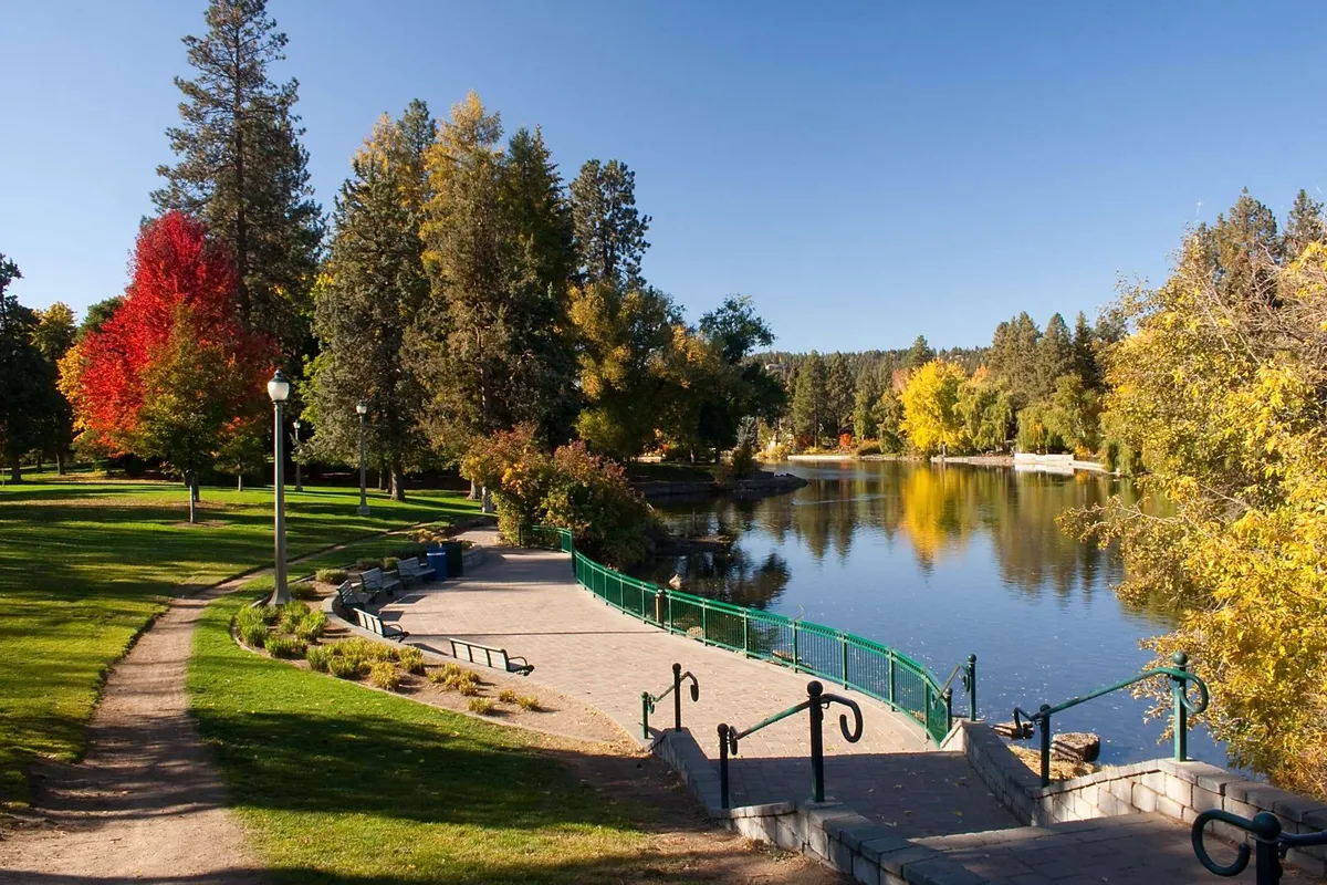A serene natural scene with calm water, colorful trees, a winding pathway, and a green railing. It suggests nearby recreational opportunities and the peaceful beauty of the surrounding area.
