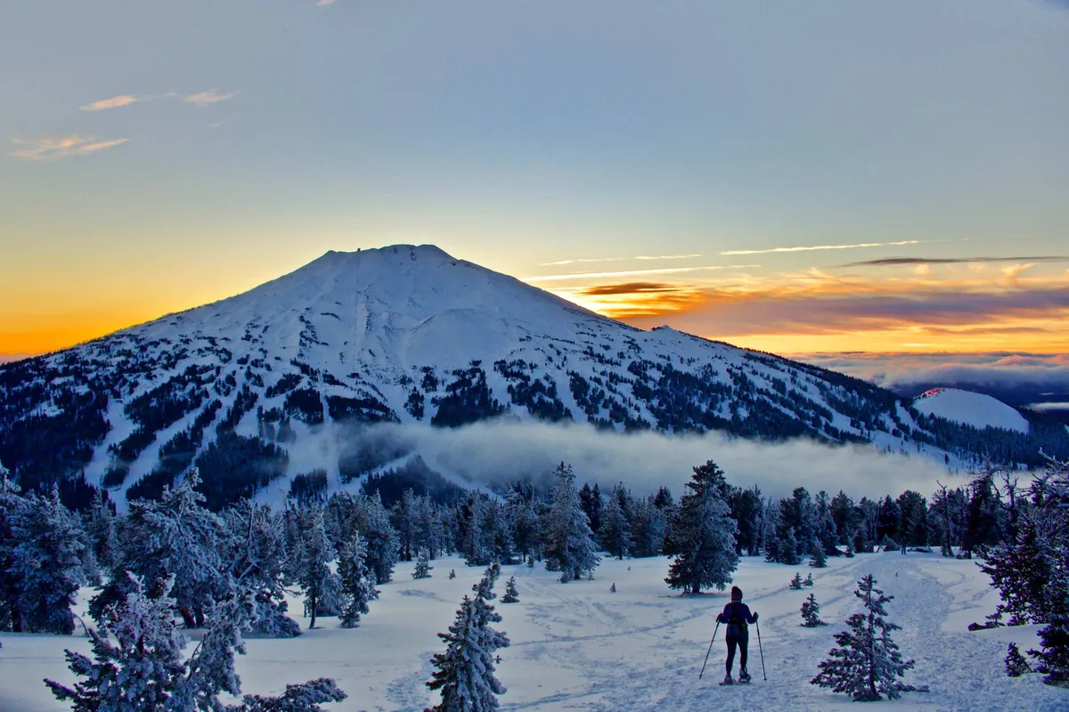 A snow-covered mountain glows under a vibrant sunset as a skier carves down the slopes, with dramatic colors in the sky and pristine white snow below. The landscape is picturesque, energetic, and highlights outdoor adventure.