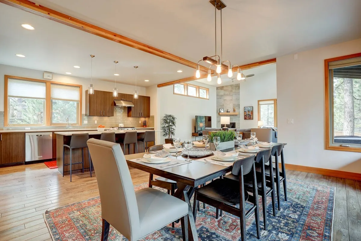 A view that includes a spacious dining table set for eight, the kitchen area behind, large windows letting in abundant light, and a decorative rug that adds warmth and style. The scene feels welcoming and designed for both everyday meals and special celebrations.