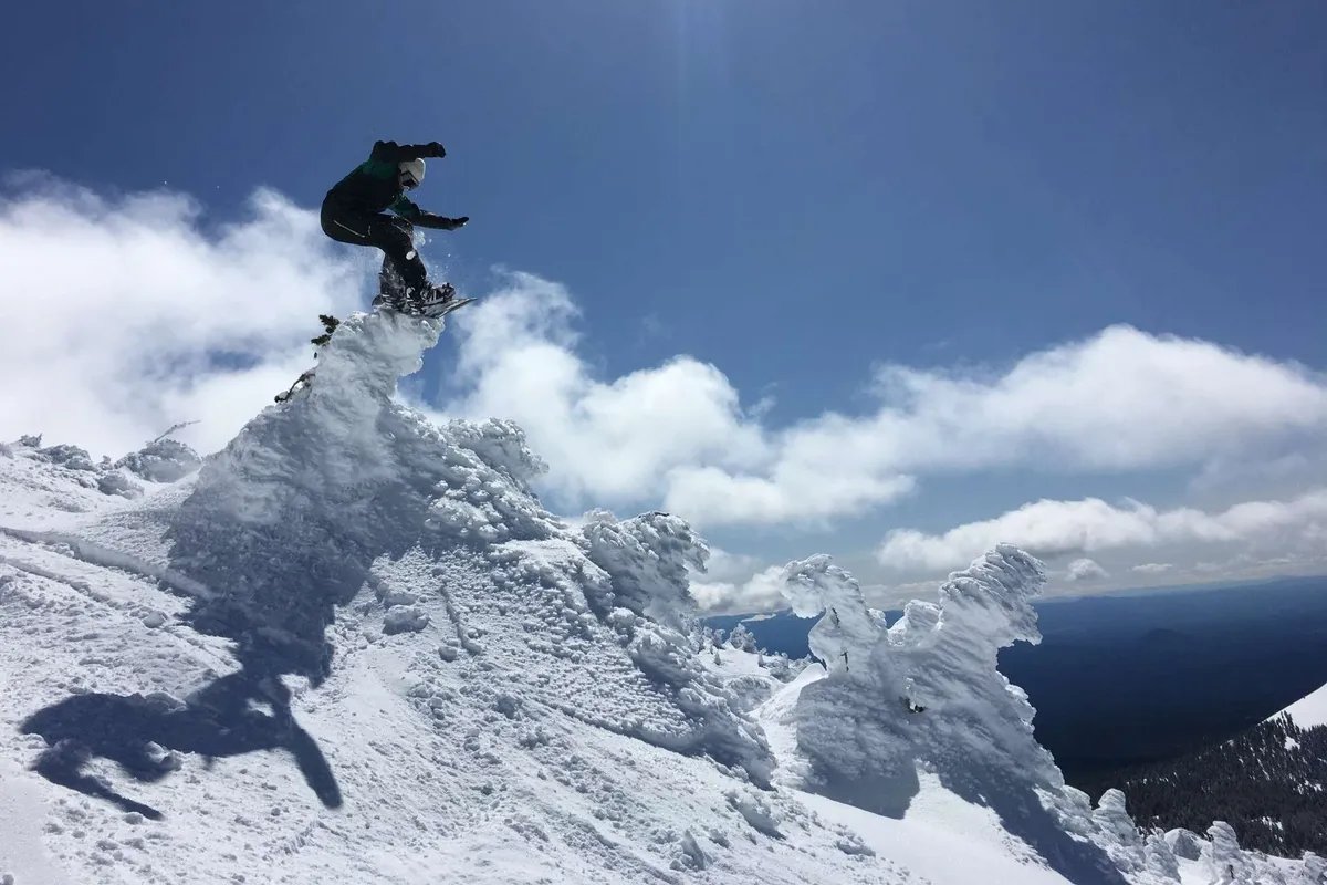 An exhilarating exterior shot of a snowboarder leaping against a backdrop of snow-covered peaks and vibrant blue skies. The scene brims with adventure, showcasing the natural beauty and recreational opportunities near Bend.