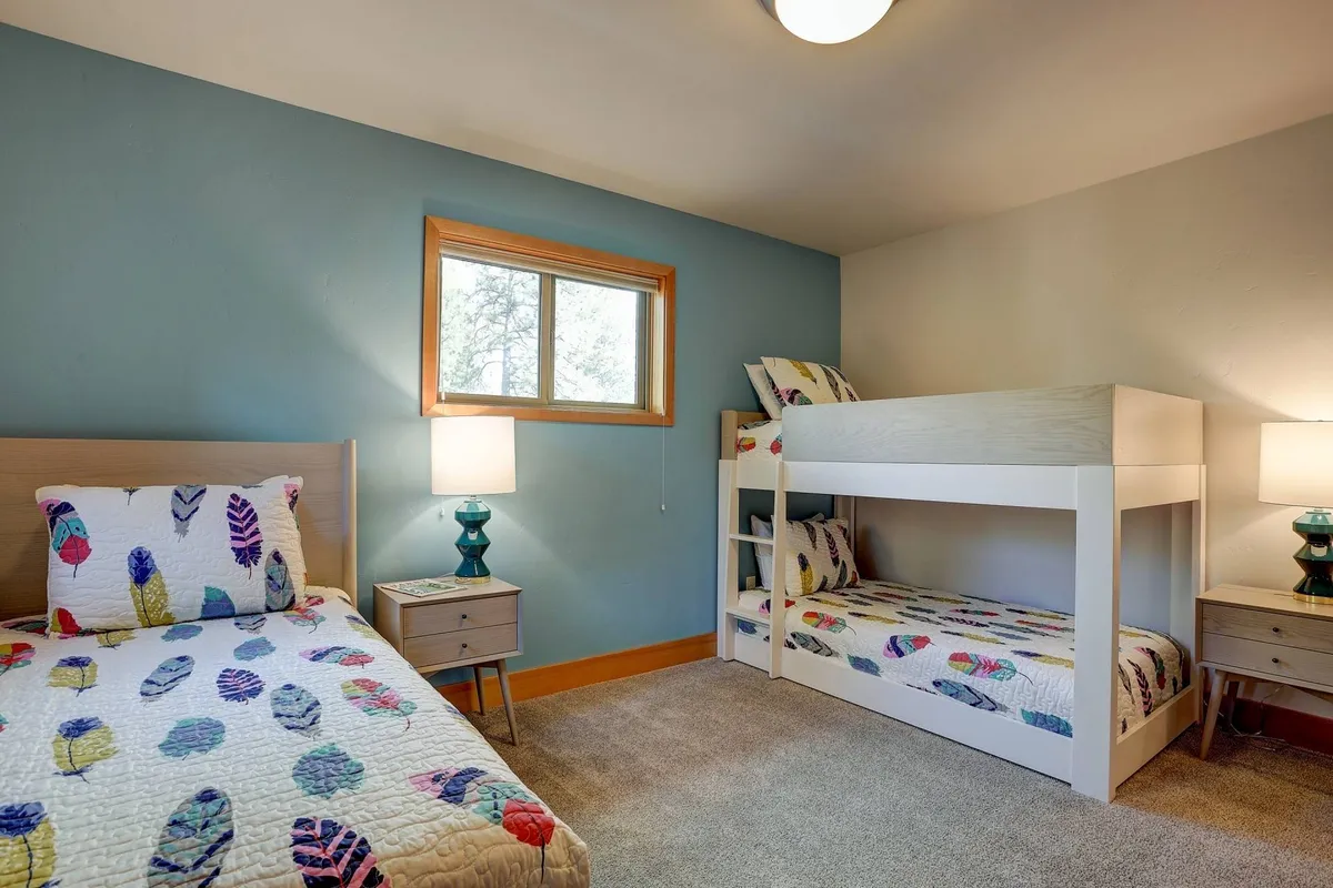 A playful and inviting bunk room with a bunk bed, single bed, cheerful nightstands with lamps, and a bright window. The space feels cozy and fun, perfect for kids or friends, and encourages both restful sleep and lighthearted moments.