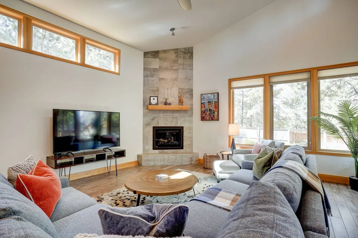 A spacious, sunlit living room featuring a large sectional sofa arranged around a modern gas fireplace. Expansive windows bathe the space in natural light, with views of the outdoors. The wooden coffee table and television complete the inviting scene, perfect for socializing, movie nights, or relaxing mornings.