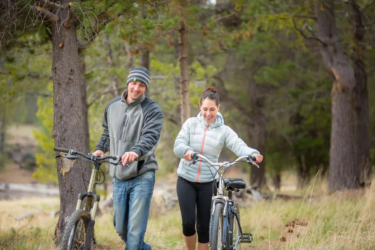 Two individuals are seen walking alongside their bicycles on a grassy path, surrounded by tall, lush trees. The open, sun-dappled area evokes a sense of freedom and natural adventure, with the bikers clearly about to embark on or returning from an outdoor excursion.