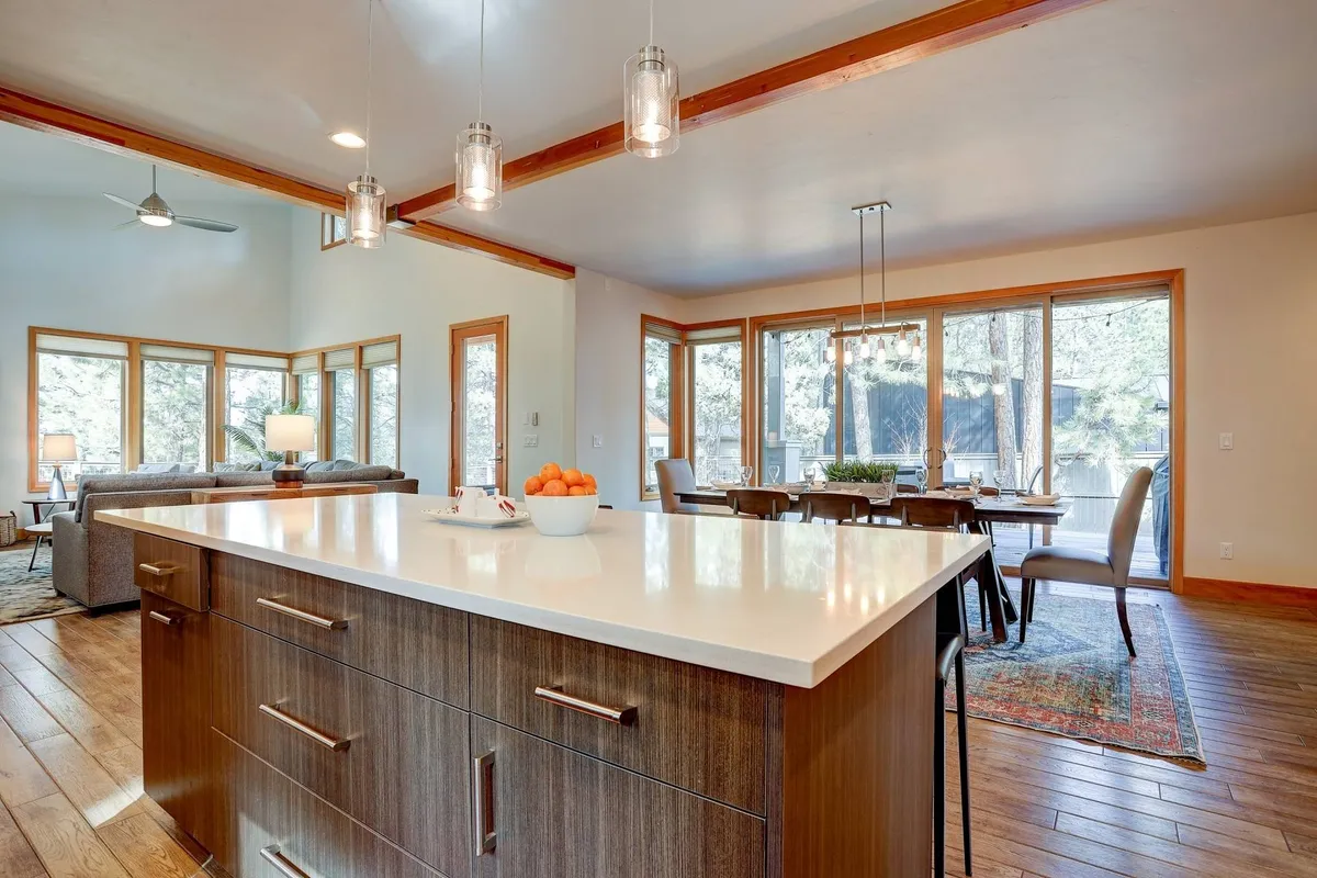 A bright, welcoming kitchen and dining area featuring a spacious island, large windows with abundant natural light, modern light fixtures, and a dining table set for gathering. The space invites shared meals and togetherness.