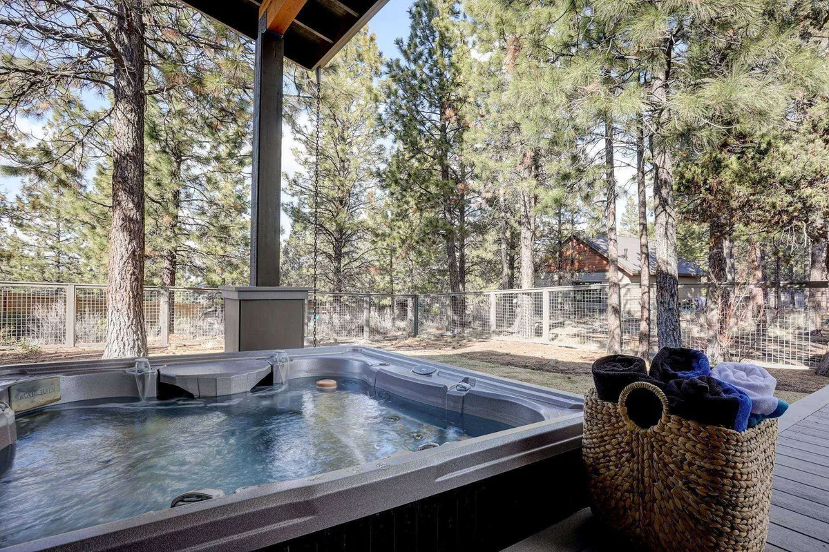 This tranquil exterior shot centers on a private, inviting hot tub surrounded by lush greenery and tall trees. In the foreground, a basket of fresh towels sits ready for guests, while the soft glow of sunset bathes the area in warmth. The sense of privacy and connection with nature is palpable, with the hot tub clearly large enough to accommodate a group.