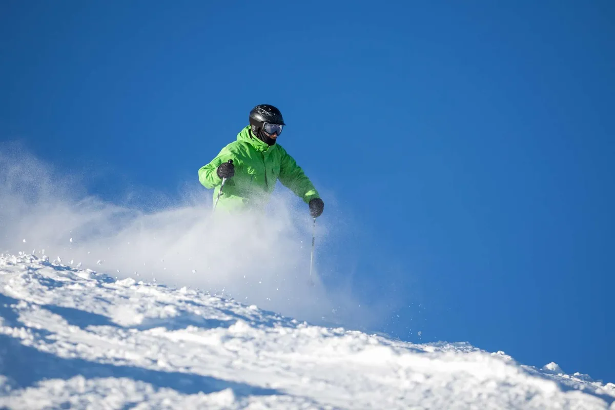 A skier carves down a pristine, snow-covered slope under a brilliant blue sky. The surrounding winter landscape suggests proximity to renowned snow sport destinations, with an atmosphere of excitement and adventure.