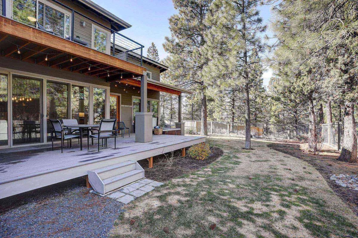A spacious deck area features a generous outdoor dining set, nestled among mature trees and overlooking landscaped pathways. The table is set for a group gathering, and string lights hint at the potential for magical evenings. The scene exudes tranquility, inviting both lively meals and quiet reflection in nature.